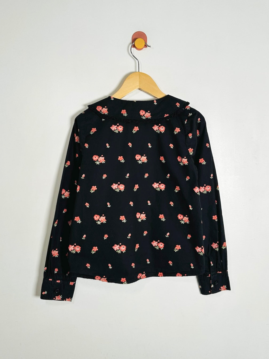 A black long-sleeve blouse with a small pink and red floral print, hanging on a wooden hanger against a plain white wall.