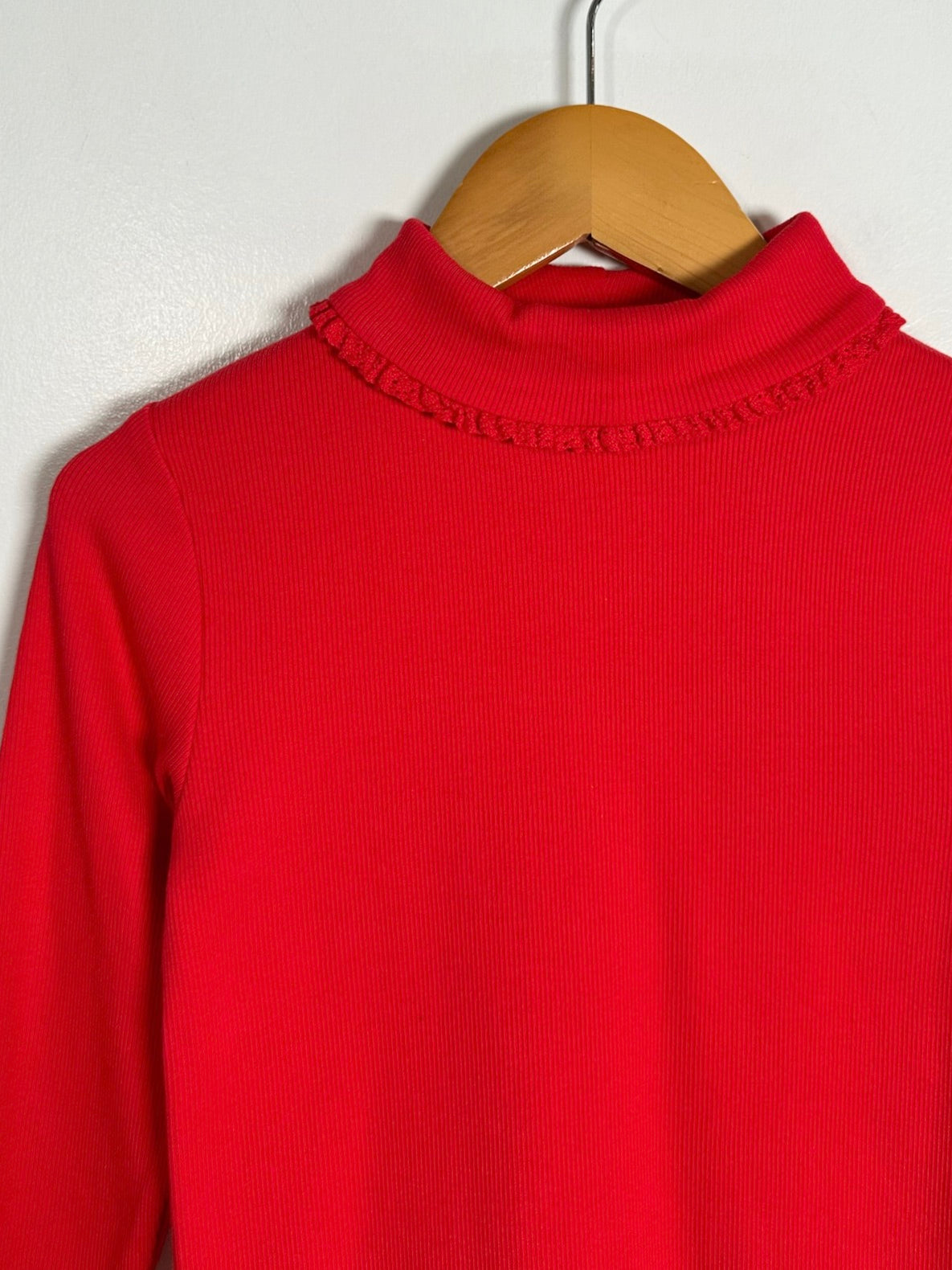 A bright red ribbed knit mock neck sweater hanging on a wooden hanger against a plain light gray wall.