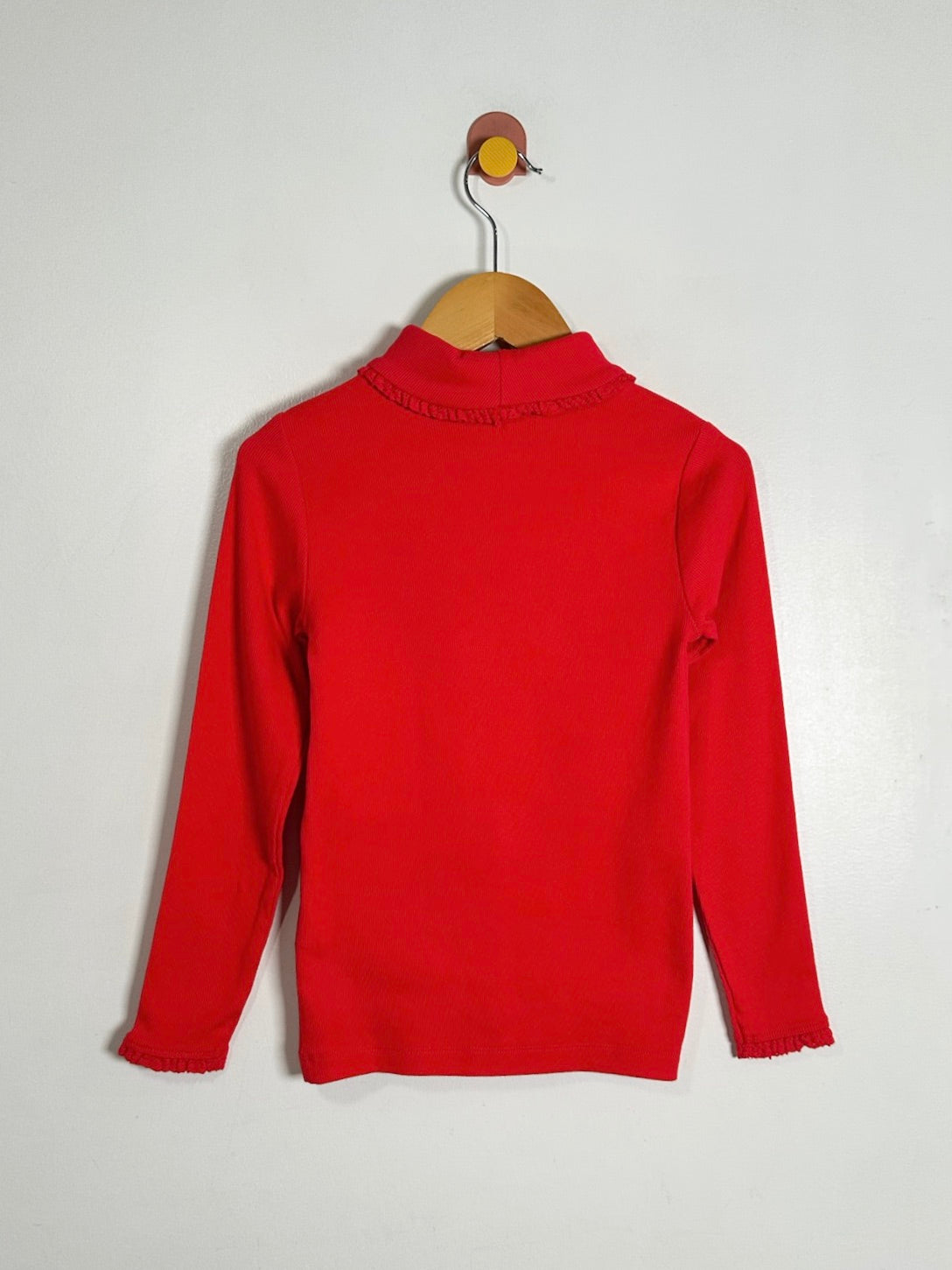 A bright red long-sleeve knit top hanging on a wooden hanger against a plain light gray wall.