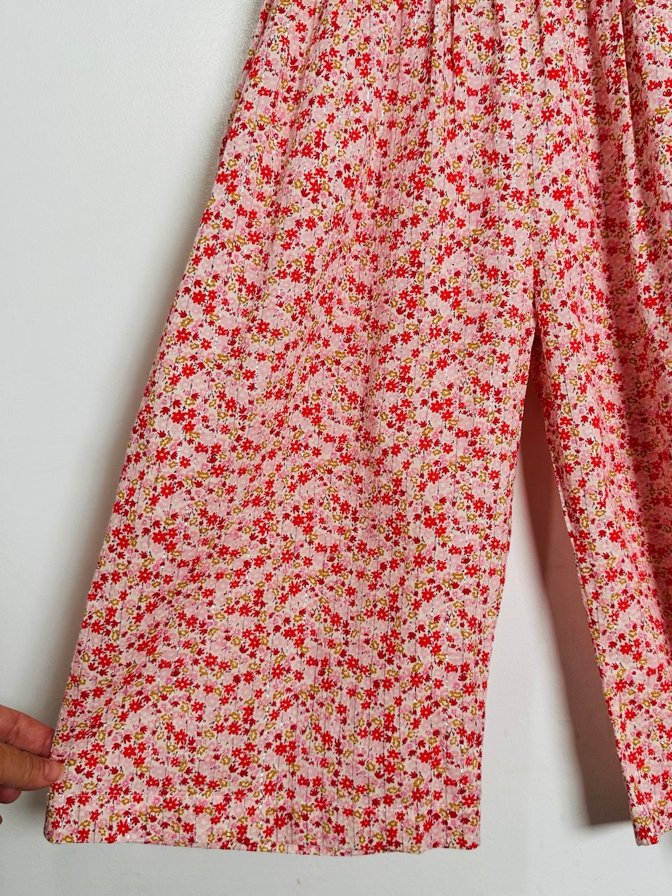 A close-up of a pair of wide-leg pants or pajama pants with a dense red and yellow floral print on a light pink background, held by a hand on the left side against a plain white wall.