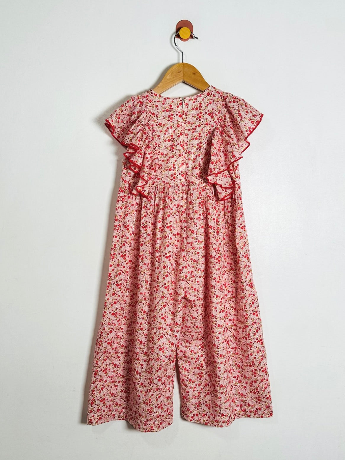 A red and white floral print jumpsuit with ruffle details on the sleeves and waist, hanging on a wooden hanger against a plain white wall.