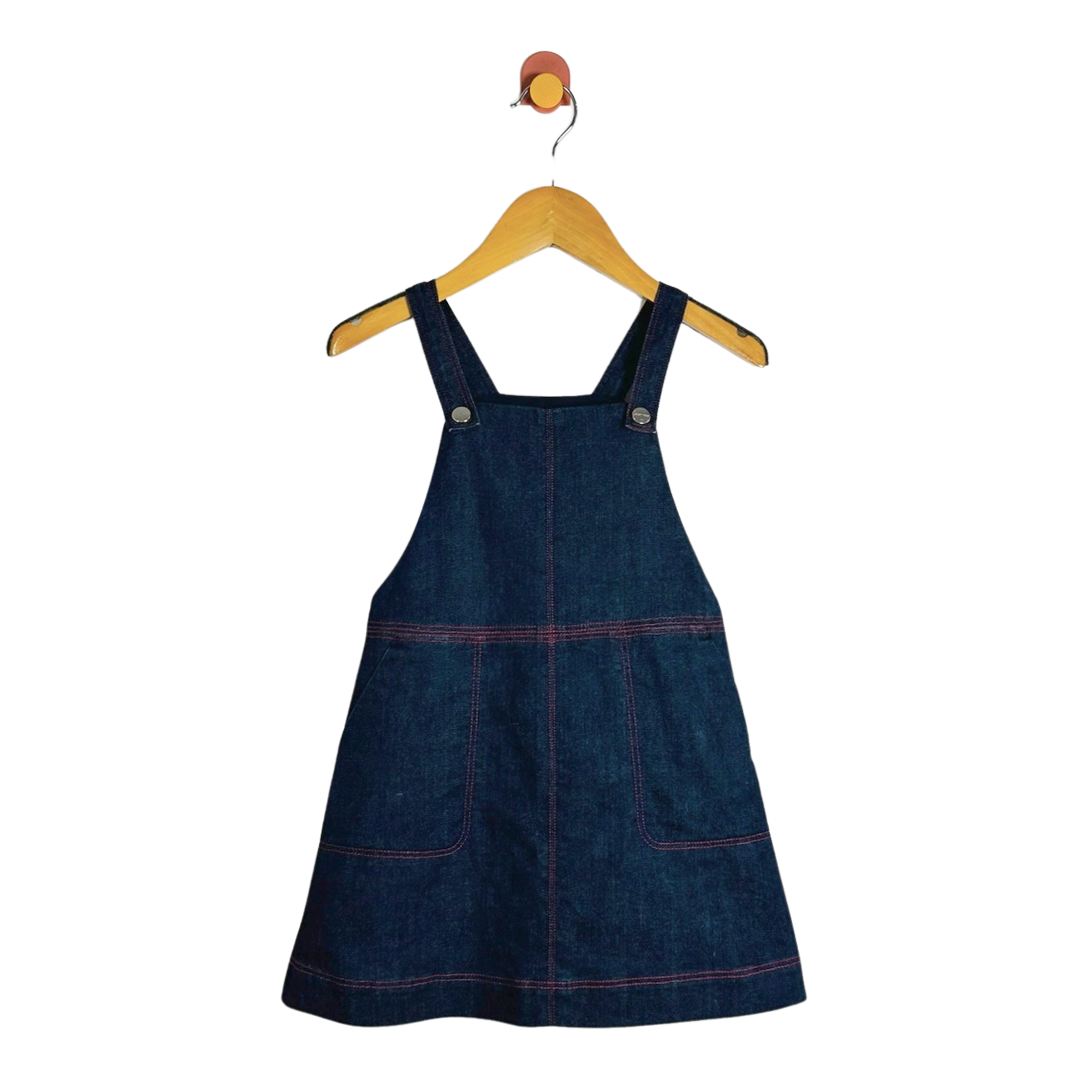A dark blue denim pinafore dress with red contrast stitching, hanging on a wooden hanger against a plain white background.