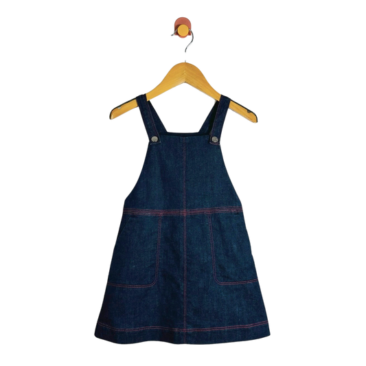 A dark blue denim pinafore dress with red contrast stitching, hanging on a wooden hanger against a plain white background.