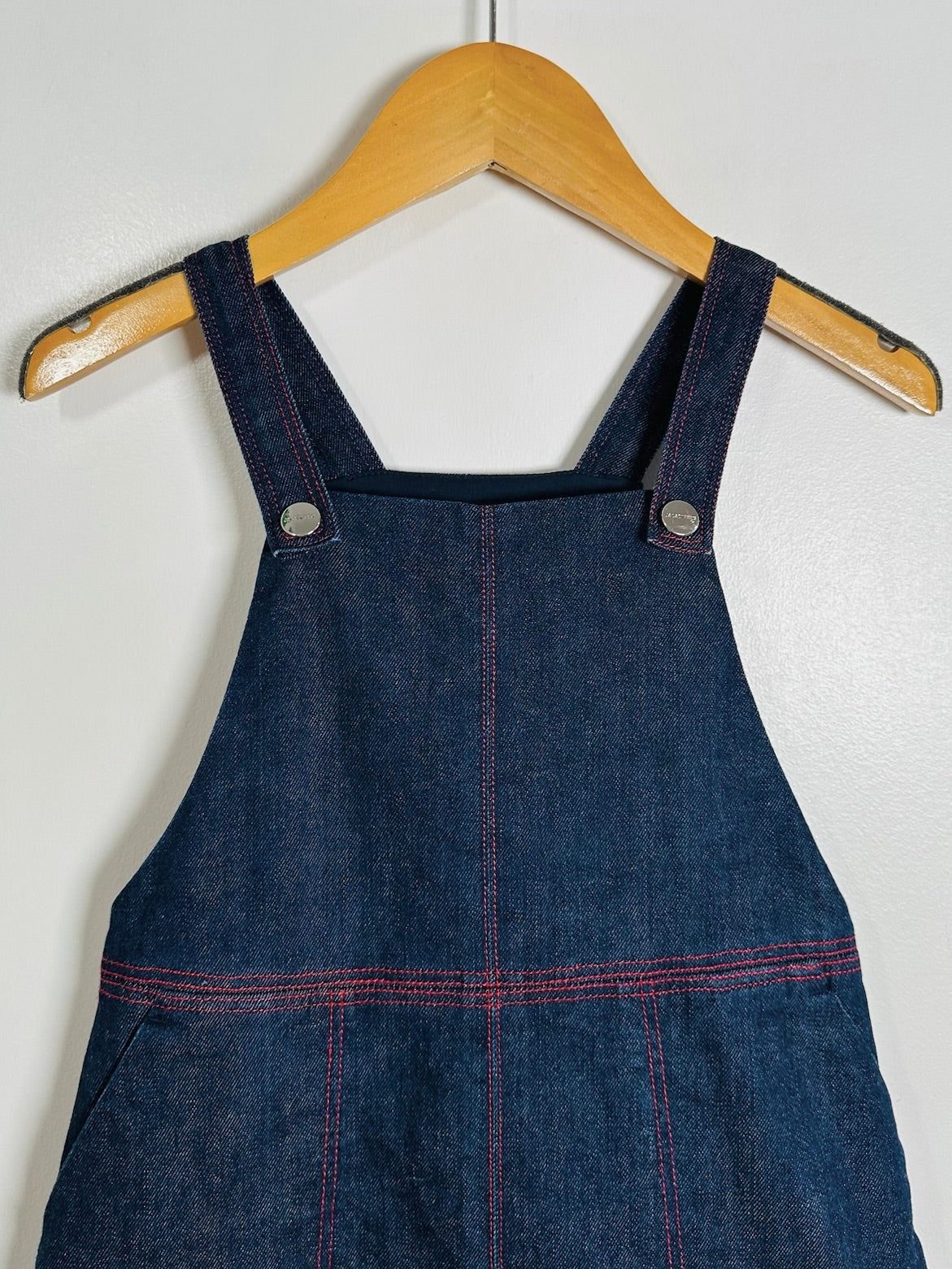 A dark blue denim bib overalls hanging on a wooden hanger against a plain light gray wall. The overalls feature red contrast stitching and silver metal snap buttons on the shoulder straps.