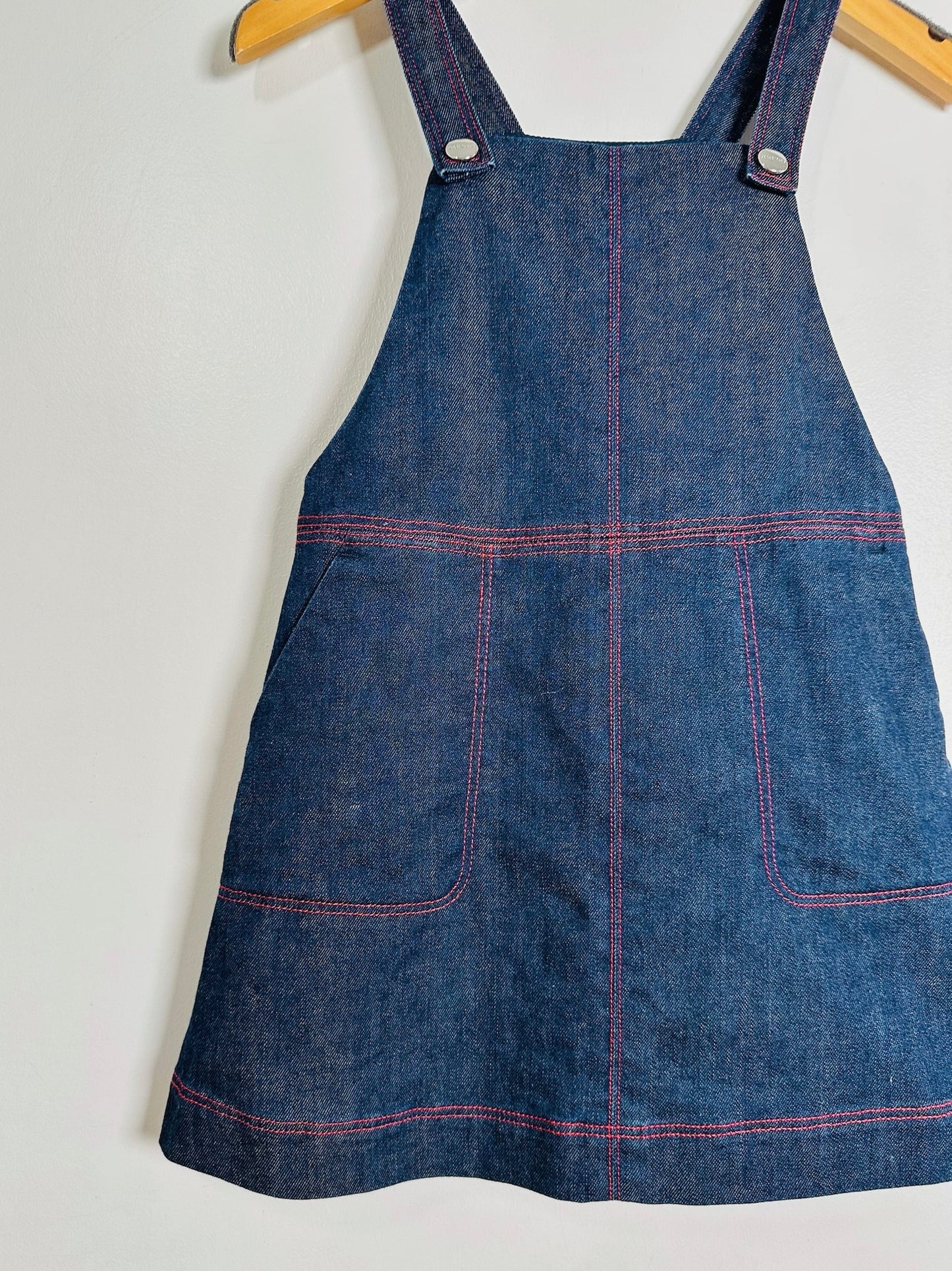 A blue denim pinafore apron with red contrast stitching, hanging on a wooden hanger against a plain white wall.
