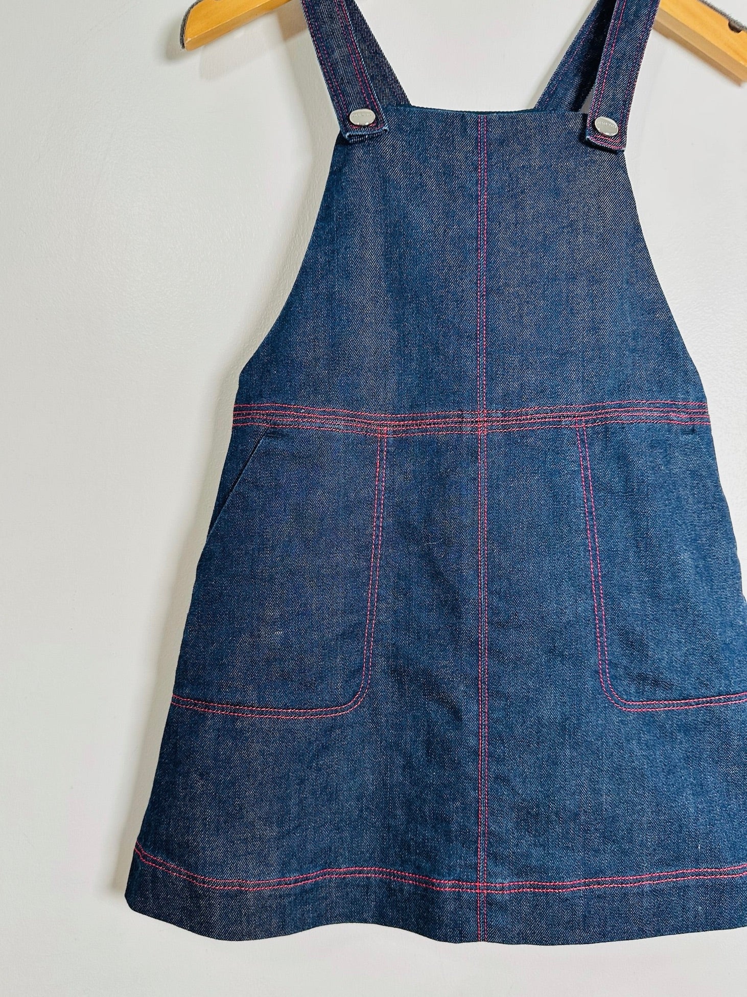A blue denim pinafore apron with red contrast stitching, hanging on a wooden hanger against a plain white wall.