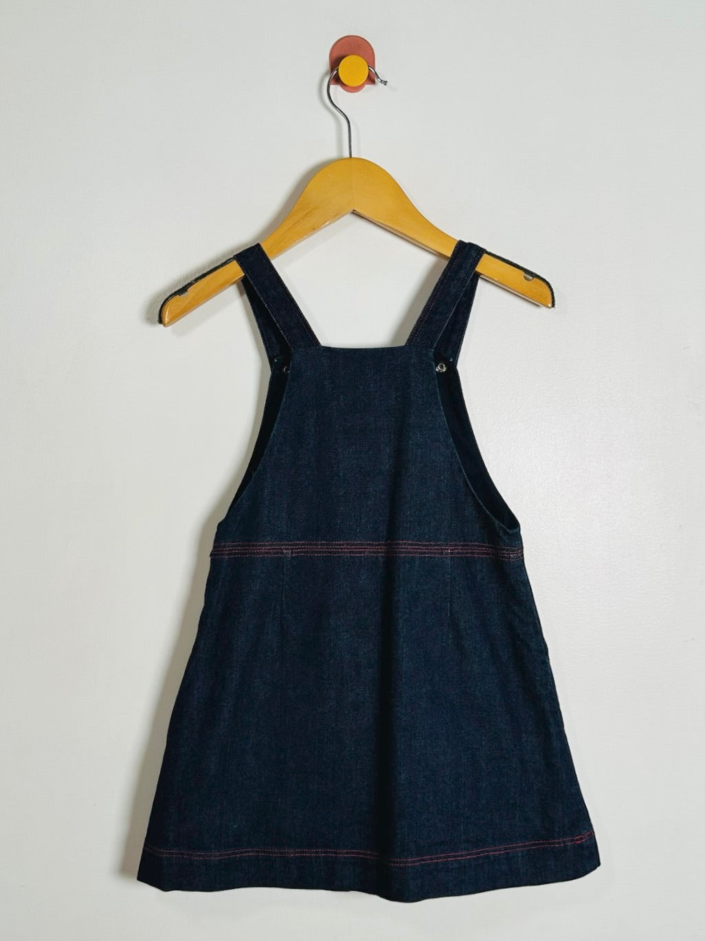 A dark indigo denim pinafore dress with red contrast stitching, hanging on a wooden hanger against a plain off-white wall.
