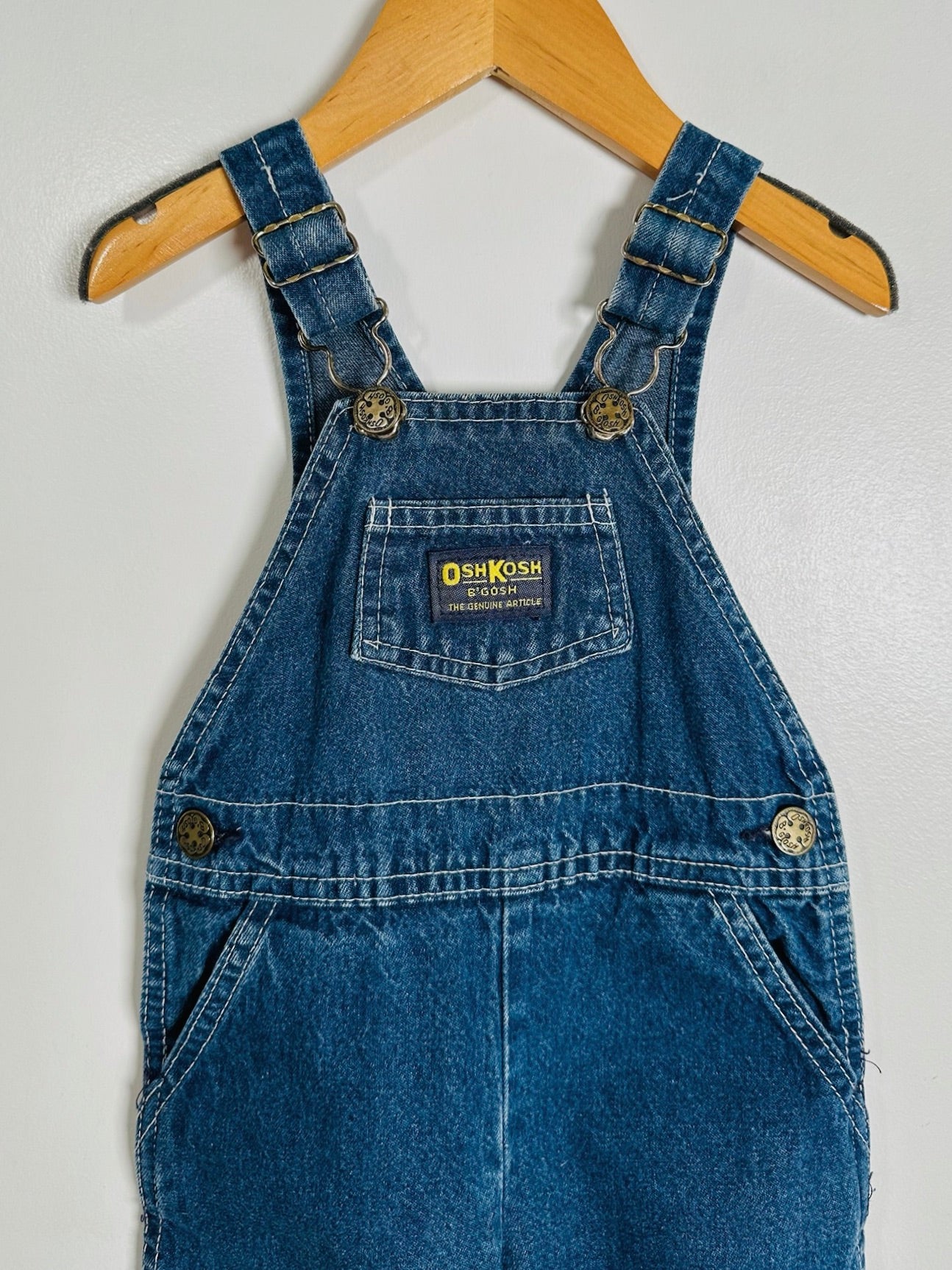 A pair of blue denim baby overalls hanging on a wooden hanger against a light gray wall. The brand label on the chest pocket reads 'Dish Kosher'.