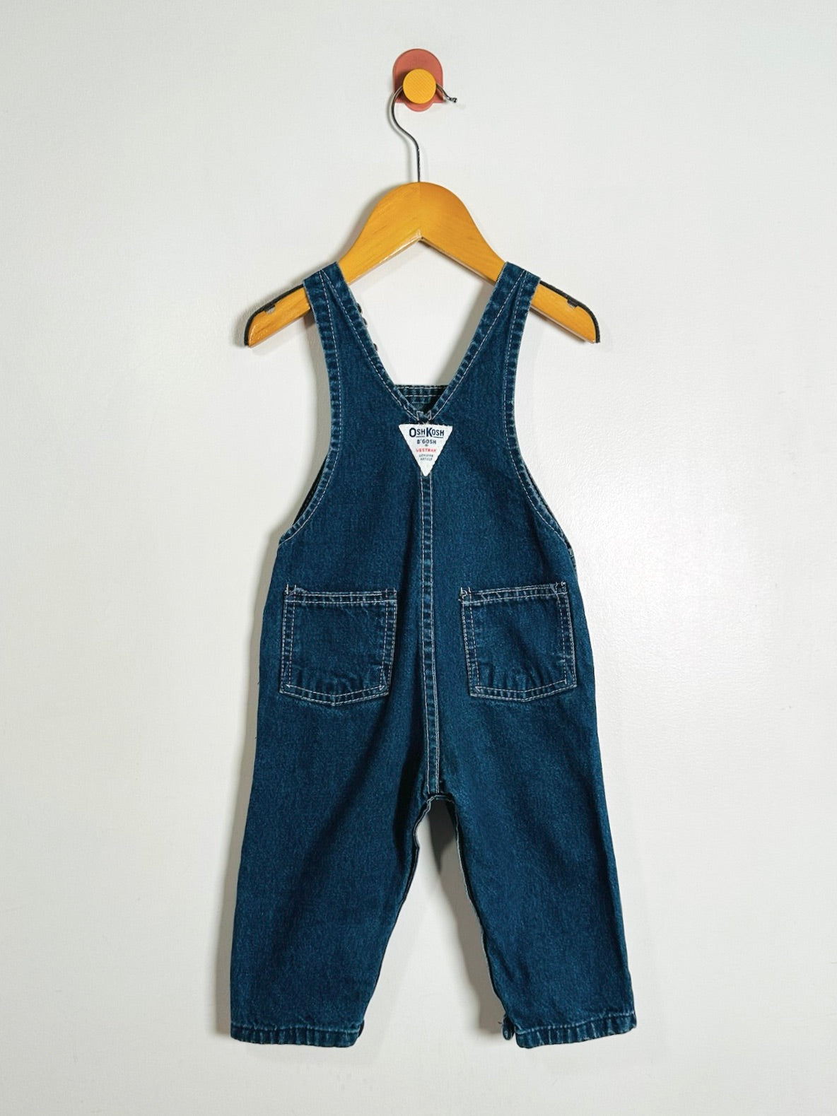 A pair of baby denim overall suspenders in dark blue, hanging from a wooden hanger on a plain white wall.