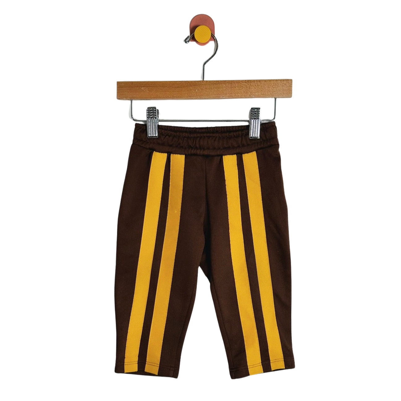 A pair of toddler brown pants with two vertical yellow stripes down the front, hanging from a wooden hanger with metal clips against a plain white background.