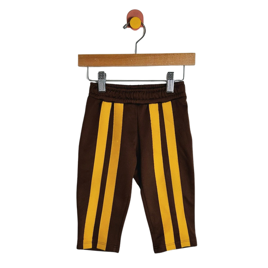 A pair of toddler brown pants with two vertical yellow stripes down the front, hanging from a wooden hanger with metal clips against a plain white background.