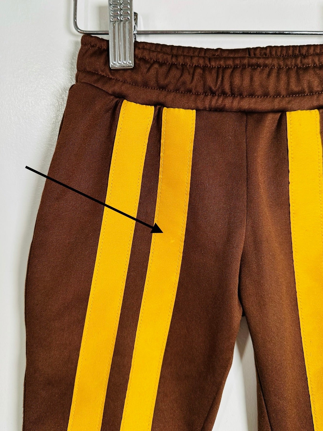 Brown pants with two wide yellow side stripes, hanging on a metal hanger against a white wall. A black arrow points to the left side of the pants.