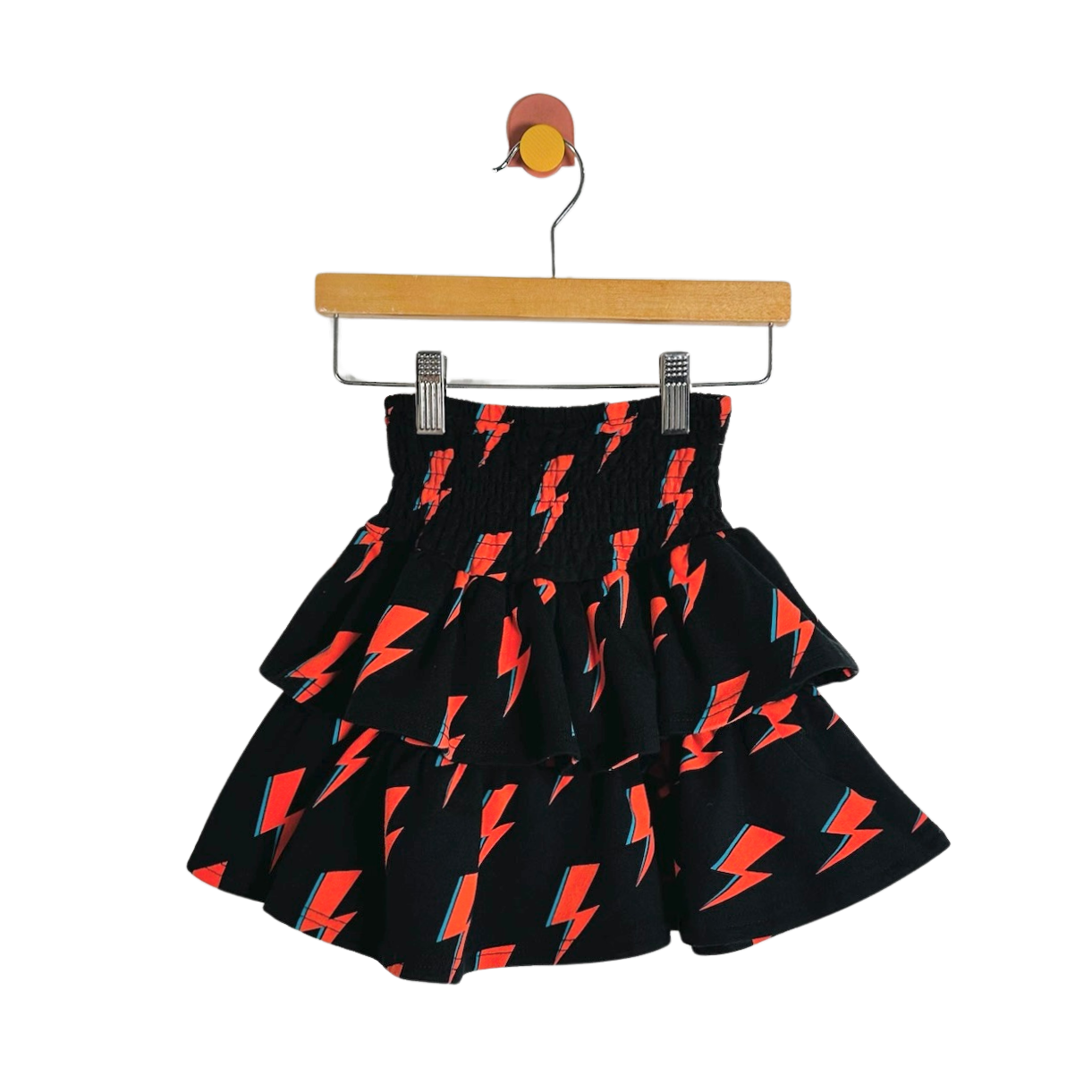 A black tiered skirt with a repeating red and orange lightning bolt pattern, hanging from a wooden hanger with metal clips against a plain white background.