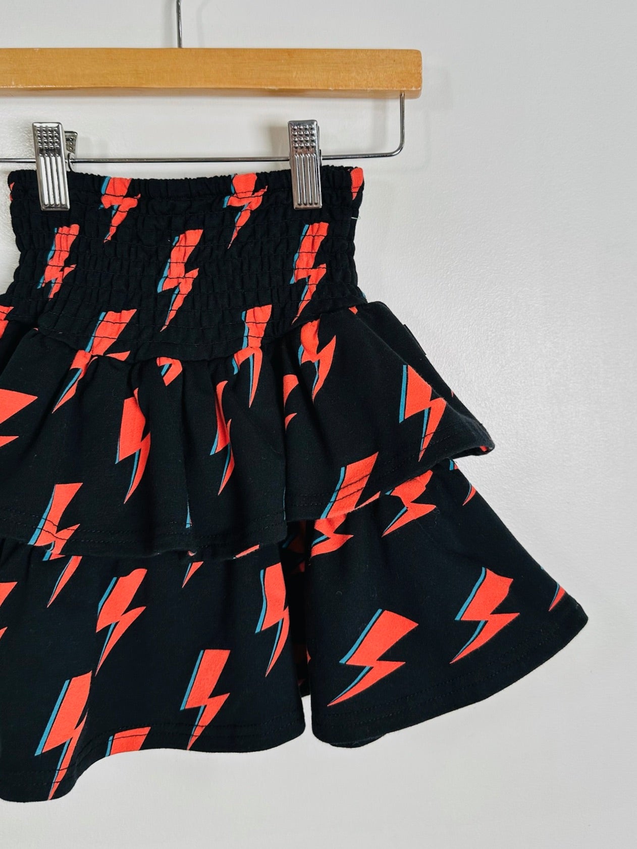 A black tiered skirt with a repeating red and orange lightning bolt pattern, hanging from a wooden hanger against a plain white wall.