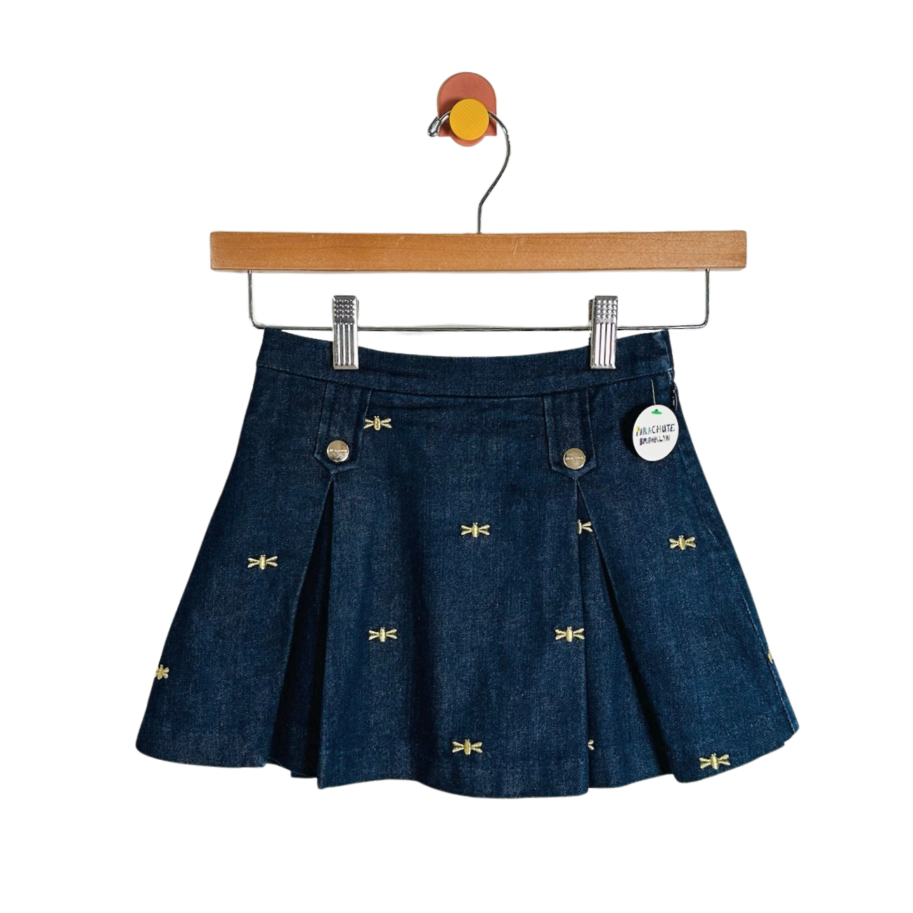 A dark blue denim pleated mini skirt with small yellow embroidered dragonflies, hanging from a wooden hanger against a white background.