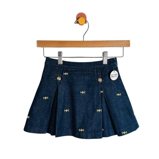 A dark blue denim pleated mini skirt with small yellow embroidered dragonflies, hanging from a wooden hanger against a white background.