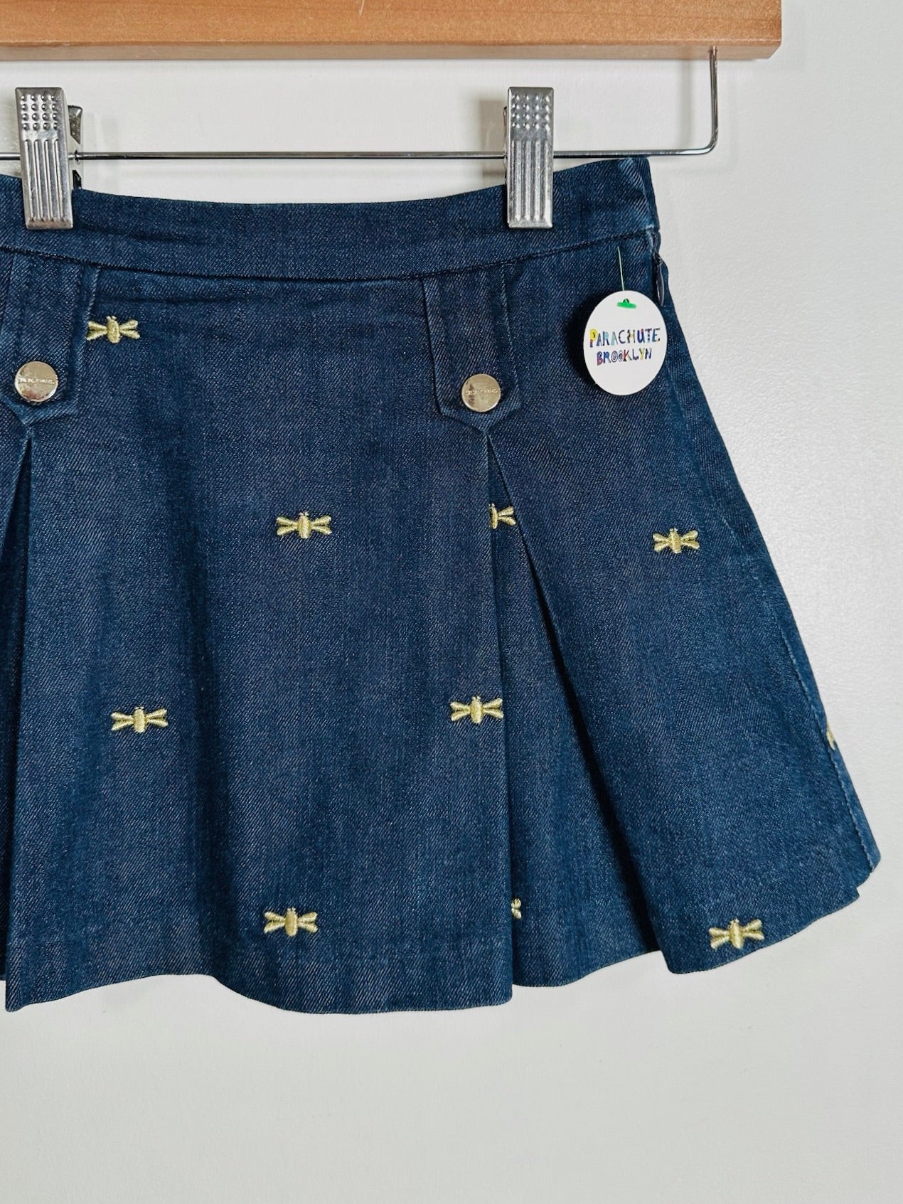 A navy blue denim skirt with small gold embroidered dragonflies, hanging from silver metal clips on a wooden hanger against a white wall. A circular paper tag is attached near the waistband.