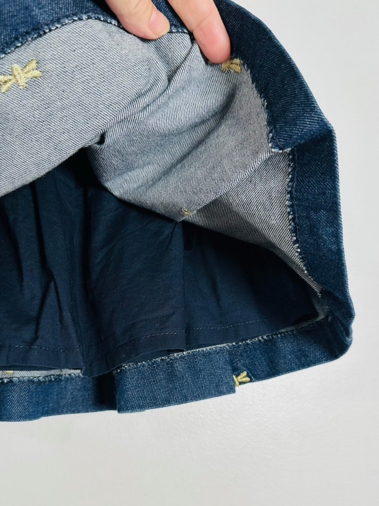 Close-up of a denim garment being held open by a hand, showing the inner lining, raw hem, and small gold embroidered logo details on a light gray background.