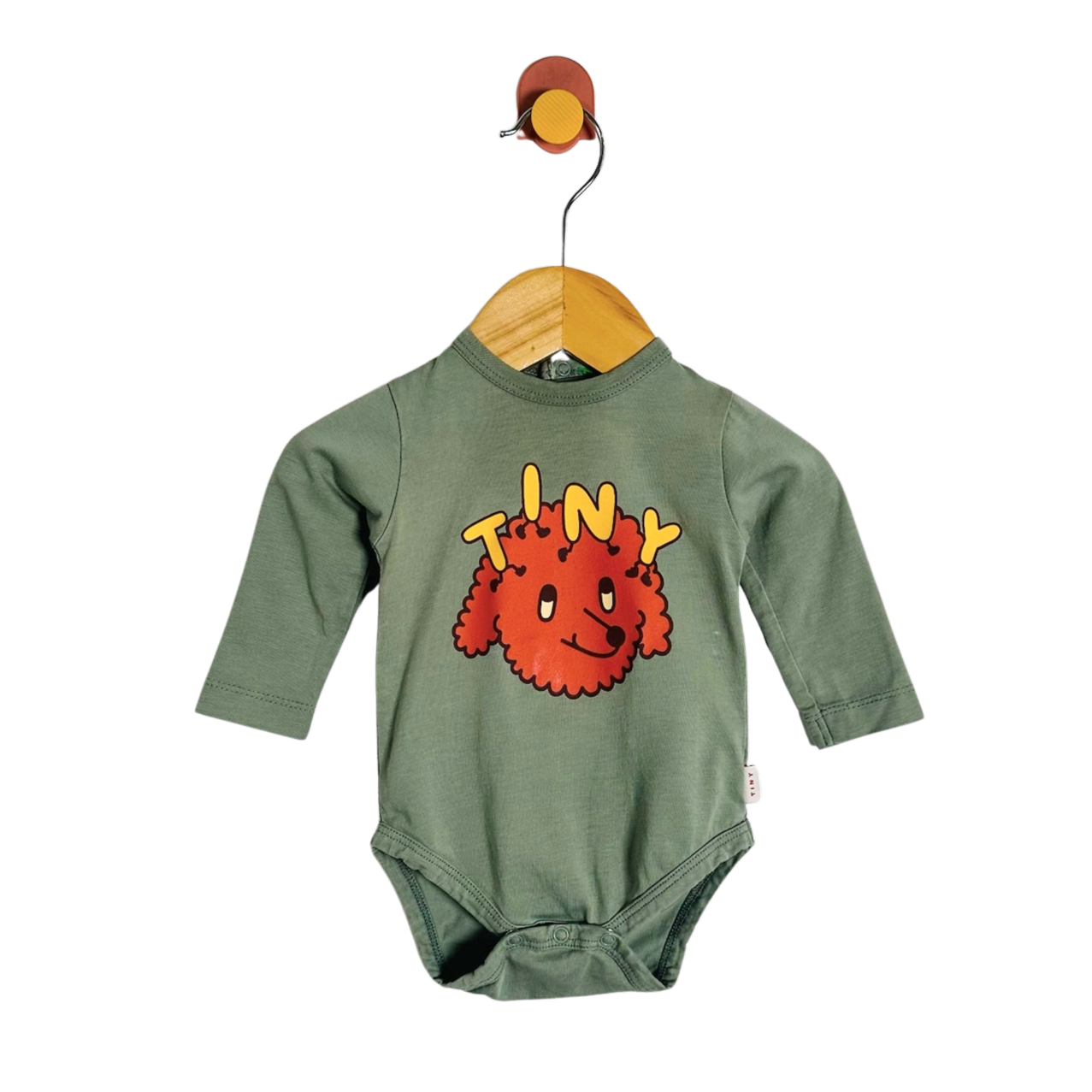A baby long-sleeve bodysuit in sage green, hanging on a wooden hanger from a wall hook. The bodysuit features a graphic of a red fuzzy cartoon character with the word 'TINY' written in yellow above it.