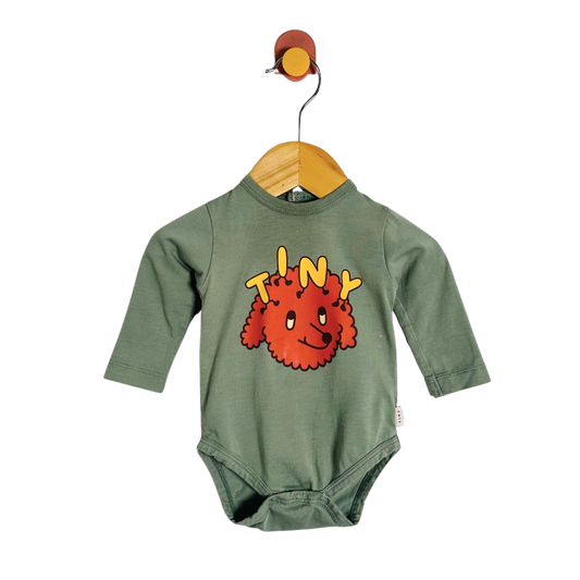 A baby long-sleeve bodysuit in sage green, hanging on a wooden hanger from a wall hook. The bodysuit features a graphic of a red fuzzy cartoon character with the word 'TINY' written in yellow above it.