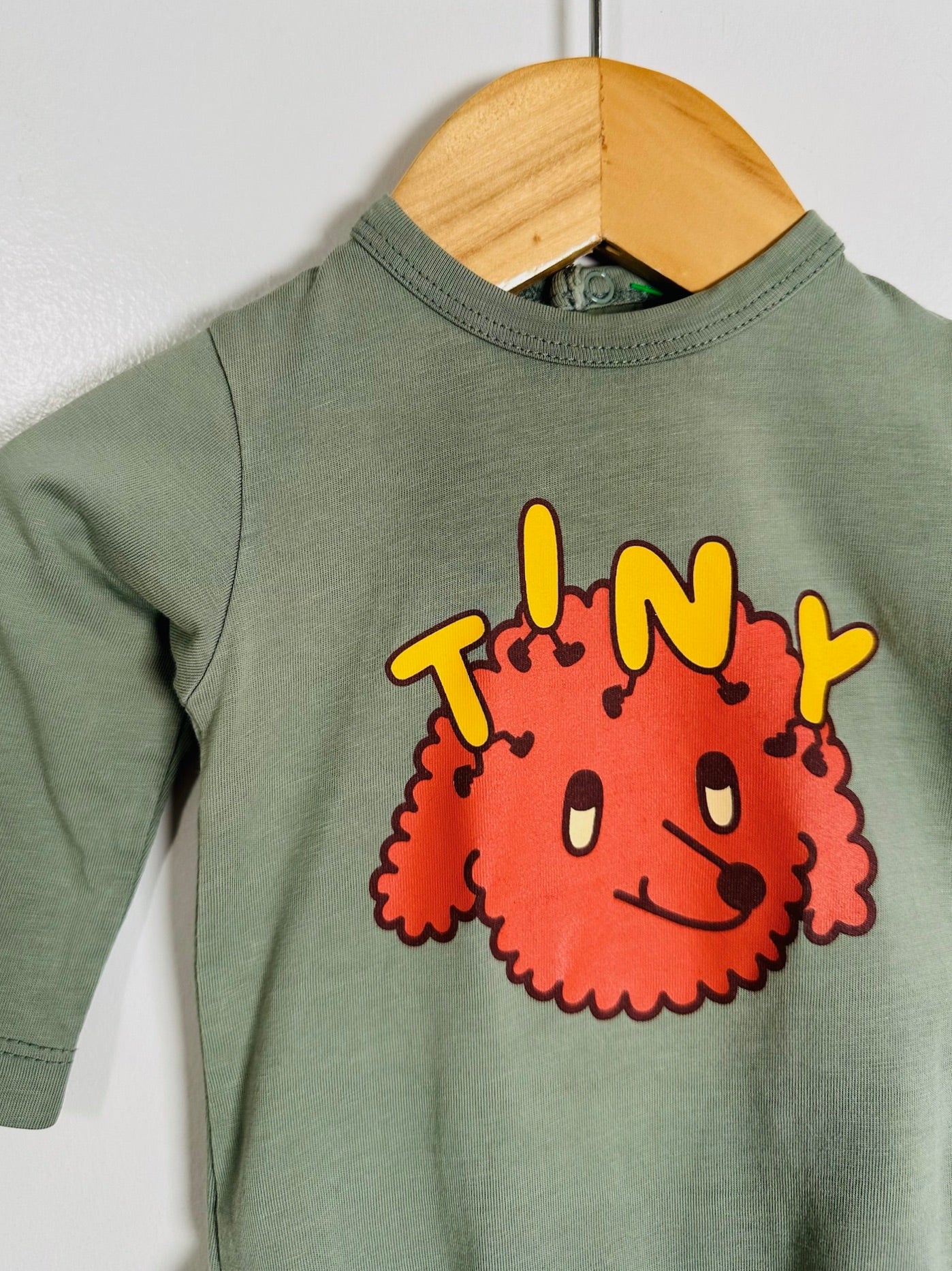 A sage green long-sleeve baby t-shirt hanging on a wooden hanger against a white wall. The shirt features a graphic of a red curly-haired cartoon dog with the word 'TINY' written in yellow above it.