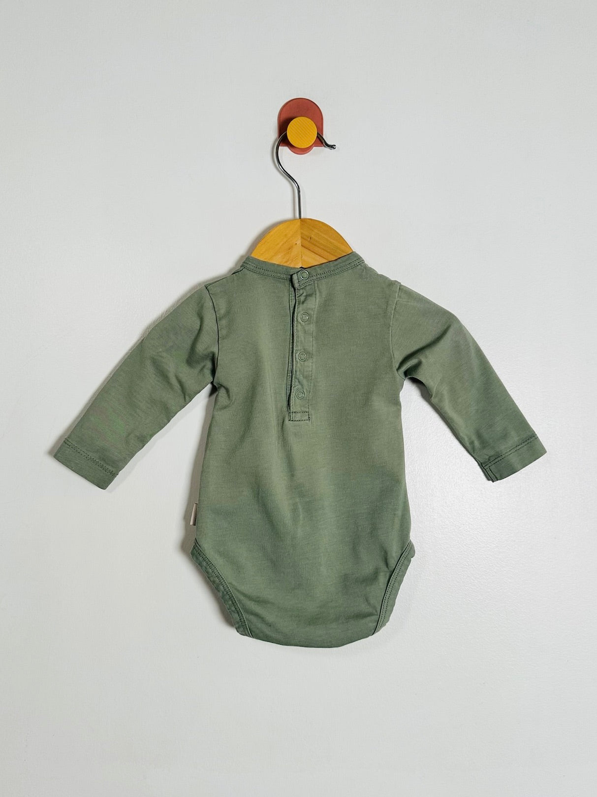 An olive green long-sleeve baby bodysuit hanging on a wooden hanger against a plain light gray wall.