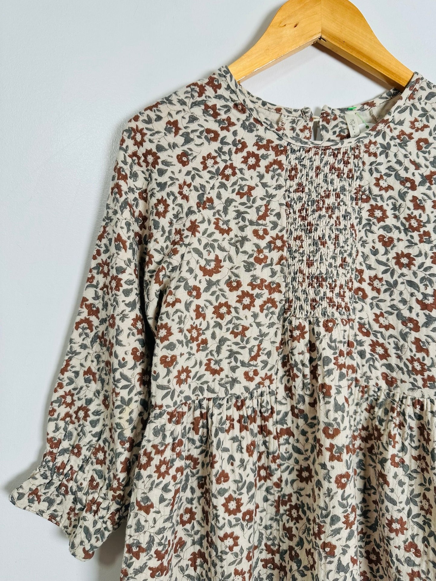 A long-sleeve tiered blouse with a dense small floral print, hanging on a wooden hanger against a plain light gray wall.