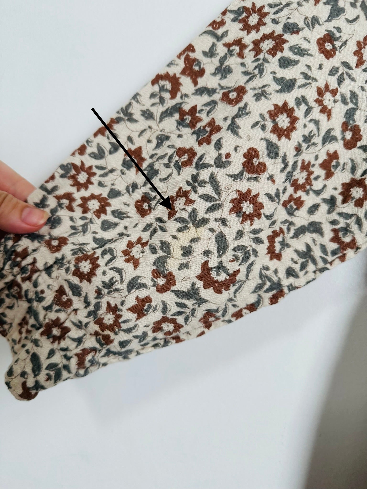 A close-up of a floral-patterned fabric sleeve or cuff being held by a hand, with a black arrow pointing to a small embroidered detail. The pattern features brown and muted blue-gray flowers with white centers on a cream background.