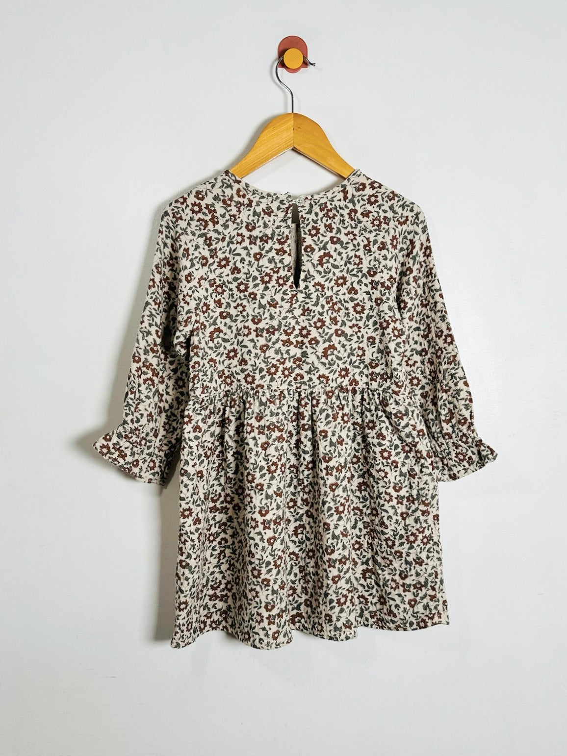 A long-sleeve babydoll dress with a brown and red floral print on a cream background, hanging on a wooden hanger against a plain white wall.