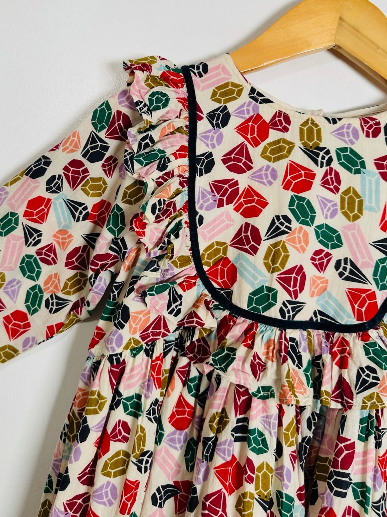 A close-up of a children's dress hanging on a wooden hanger against a plain white wall. The dress has a colorful geometric gemstone print on a cream background with black trim around the neckline.