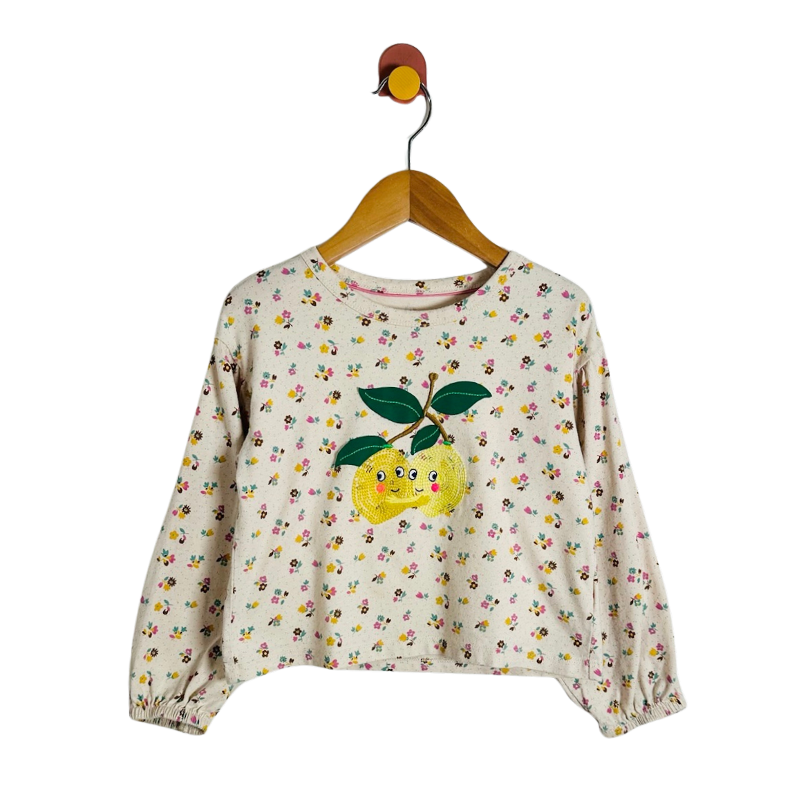 A cream-colored long-sleeve crop top with a dense yellow, pink, and green floral print, featuring a large embroidered yellow lemon with green leaves on the front. The top is displayed on a wooden hanger against a solid white background.