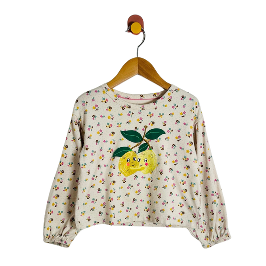 A cream-colored long-sleeve crop top with a dense yellow, pink, and green floral print, featuring a large embroidered yellow lemon with green leaves on the front. The top is displayed on a wooden hanger against a solid white background.
