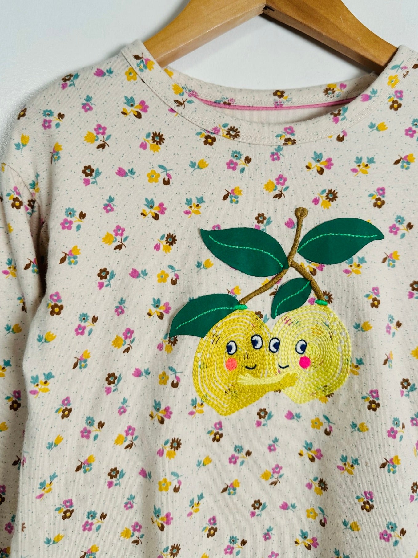 A cream-colored long-sleeve sweatshirt with a dense yellow, pink, and blue floral print, featuring a large applique of two smiling yellow lemons with green leaves on the front, hanging on a wooden hanger.