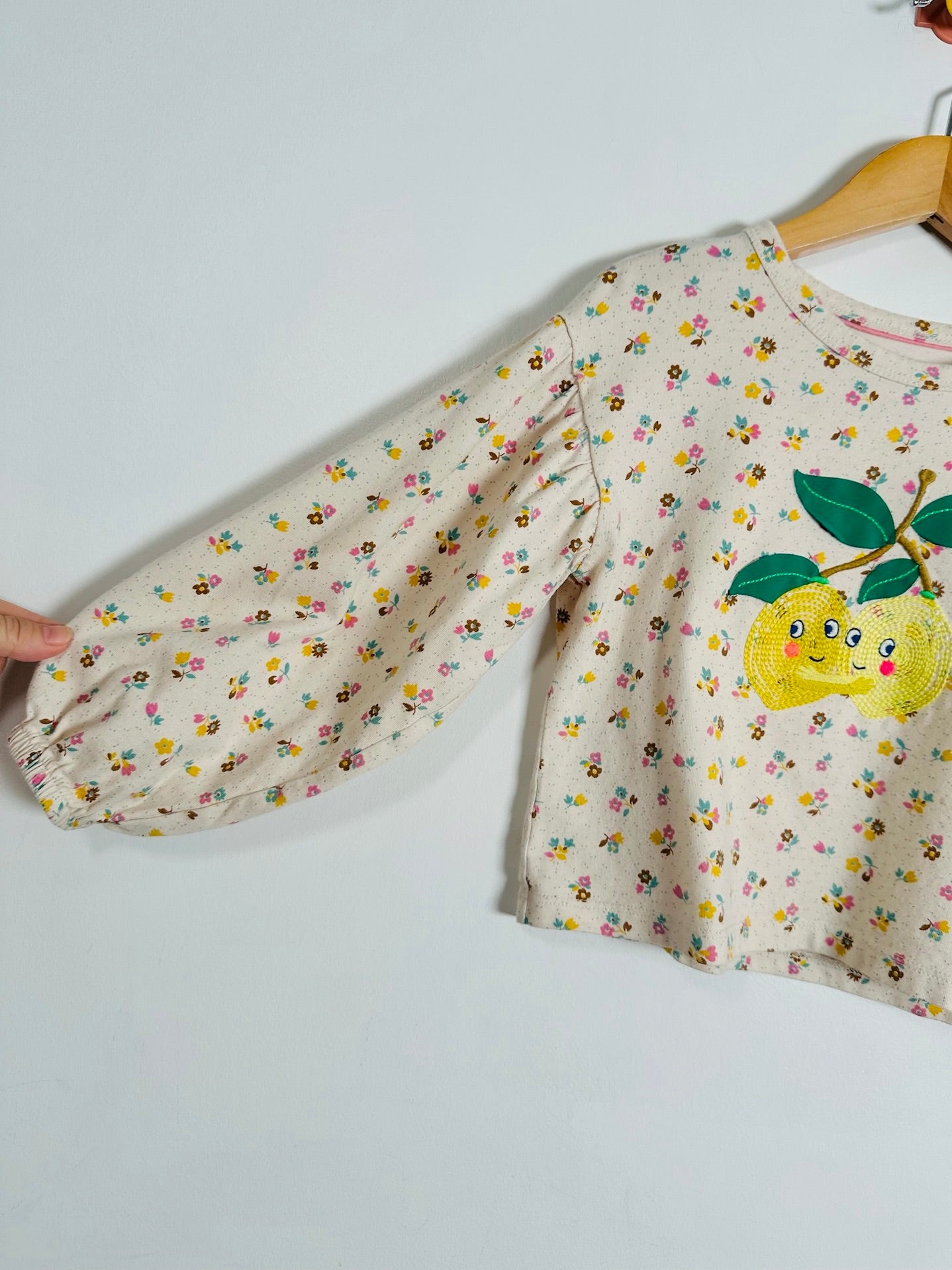 A children's long-sleeve top hanging on a wooden hanger against a plain light background. The shirt has a cream base with a dense multicolor floral print and a large embroidered yellow lemon character on the chest with green leaves.