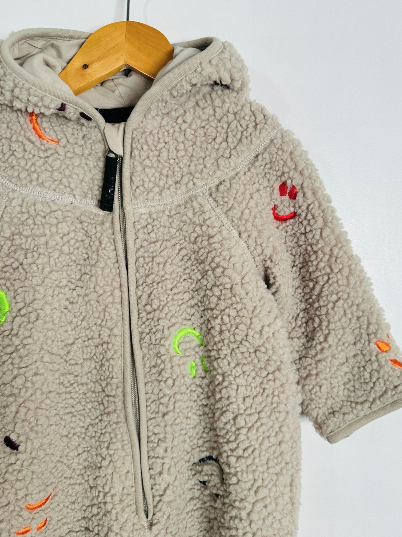 A beige sherpa fleece zip-up hoodie with colorful embroidered smiley faces, hanging on a wooden hanger against a plain white background.