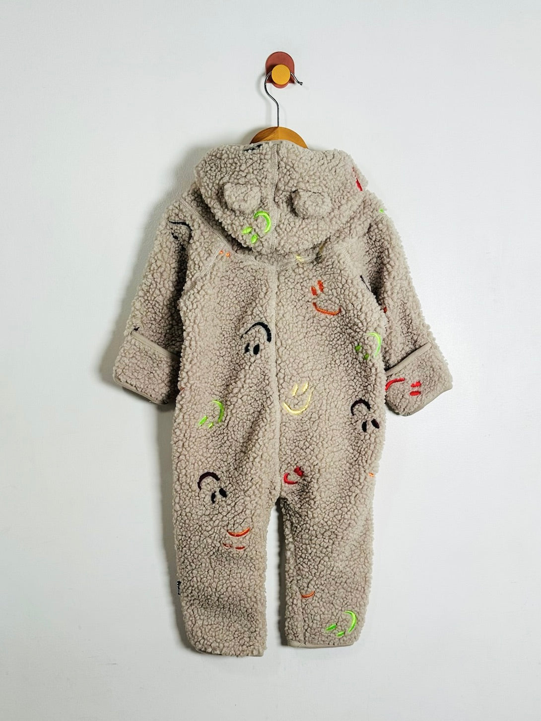 A baby's fleece hooded jumpsuit in a warm taupe-beige color, decorated with colorful embroidered faces and smiley faces, hanging on a wooden hanger against a plain white wall.
