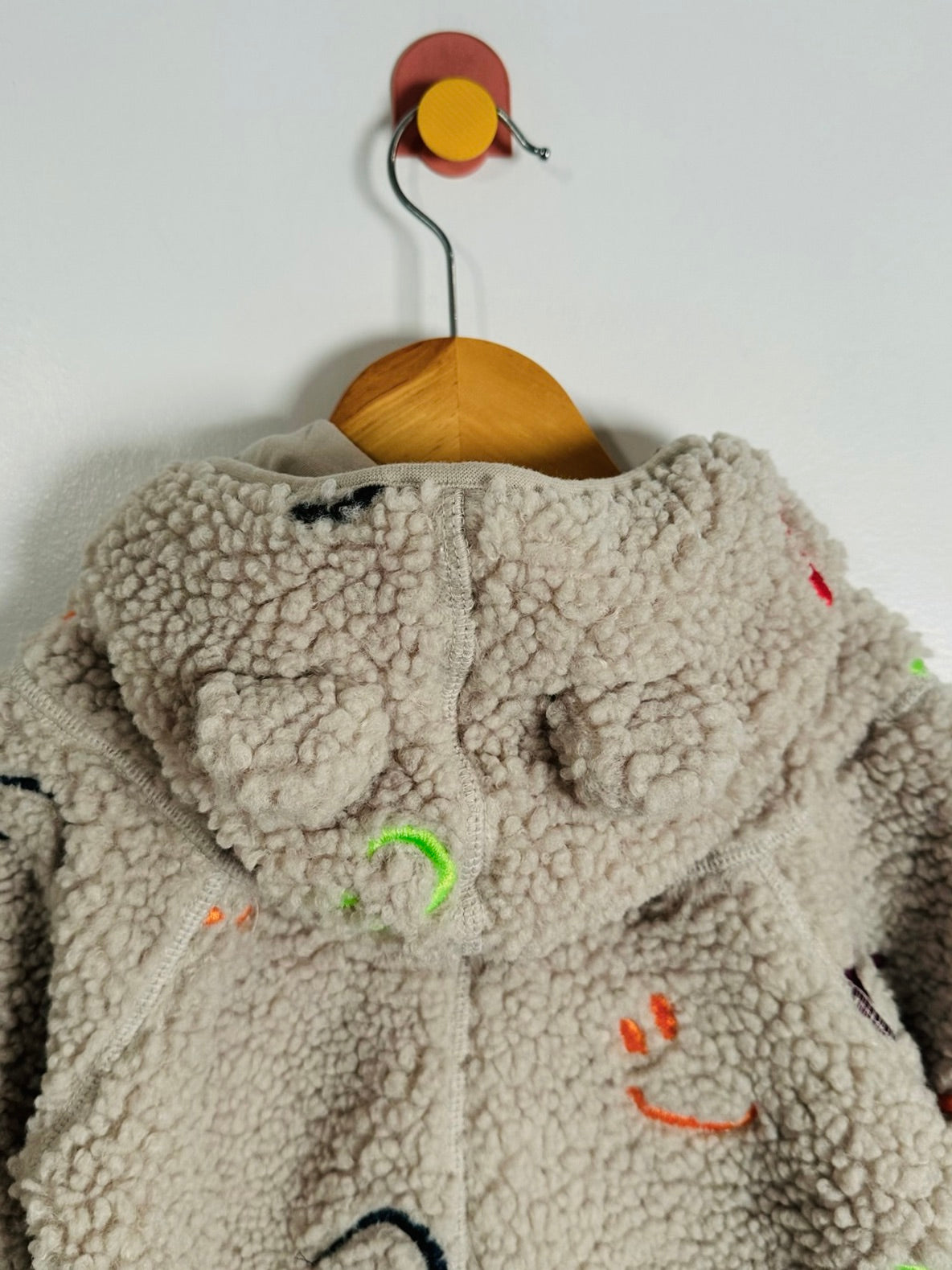 A beige sherpa fleece hooded jacket with colorful embroidered smiley faces, hanging on a wooden hanger against a plain white wall.