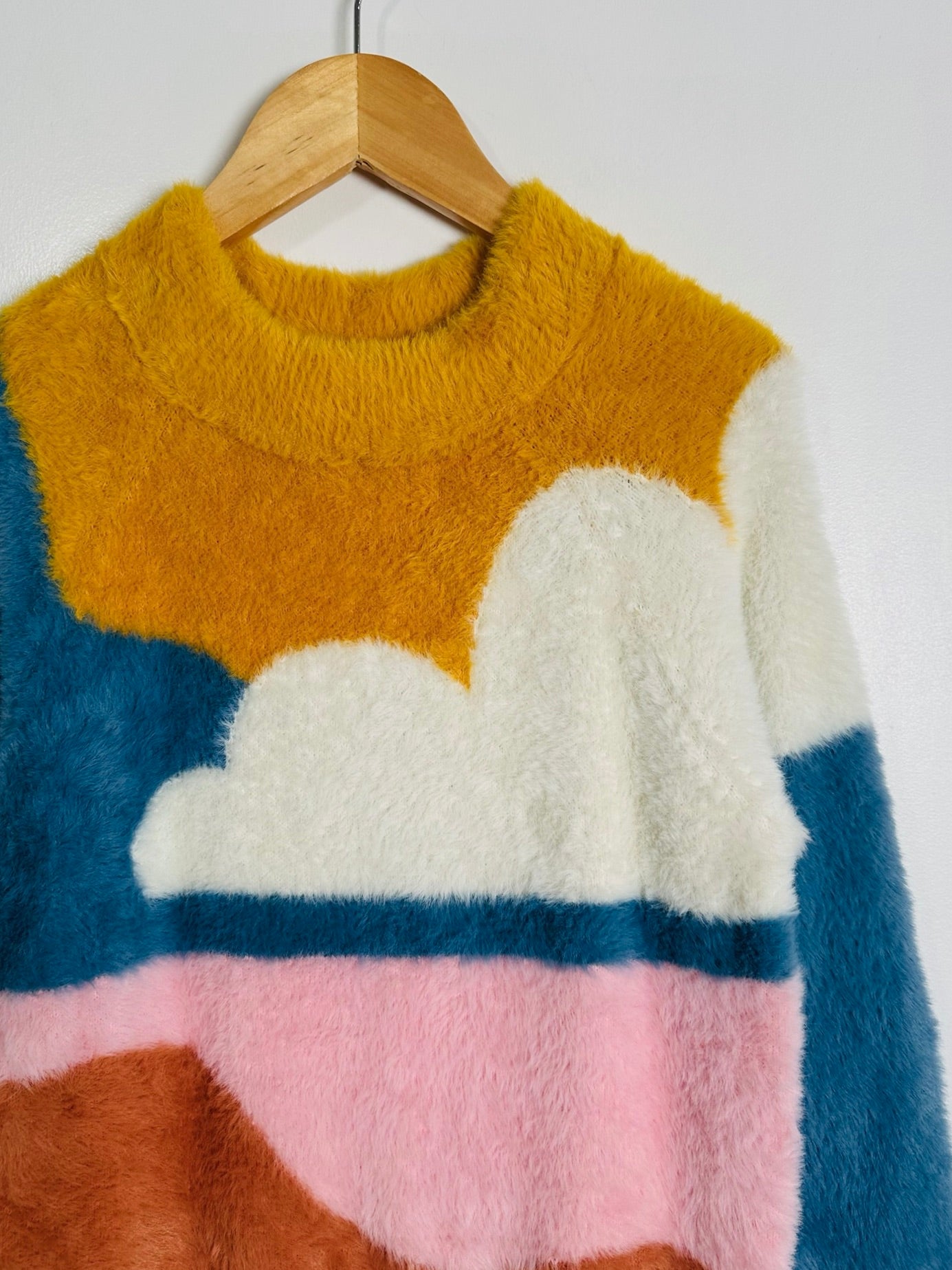 A fuzzy knit sweater hanging on a wooden hanger against a plain light background. The sweater has a color-blocked cloud pattern with mustard yellow, white, blue, pink, and brown sections.