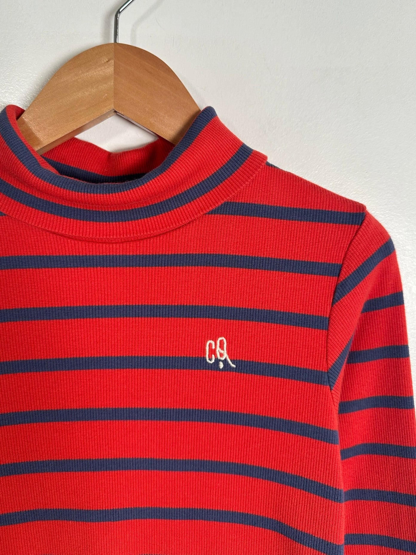 A red ribbed mock neck sweater with navy blue horizontal stripes, hanging on a wooden hanger against a plain off-white wall. A small embroidered 'C&L' logo is visible on the left chest.