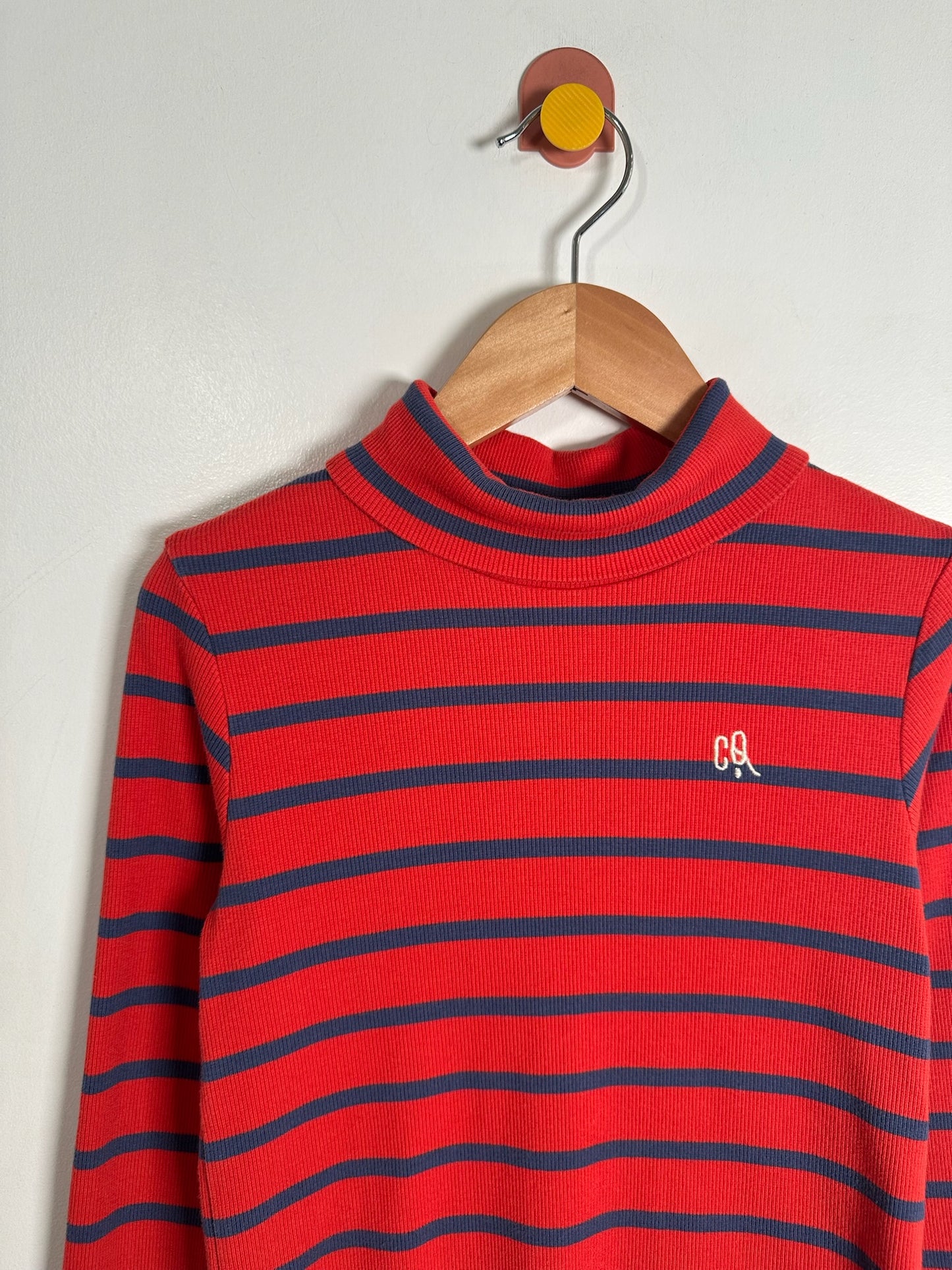 A red and navy blue horizontally striped mock neck sweater hanging on a wooden hanger against a plain white wall. A small white embroidered 'CR' logo is visible on the left chest.