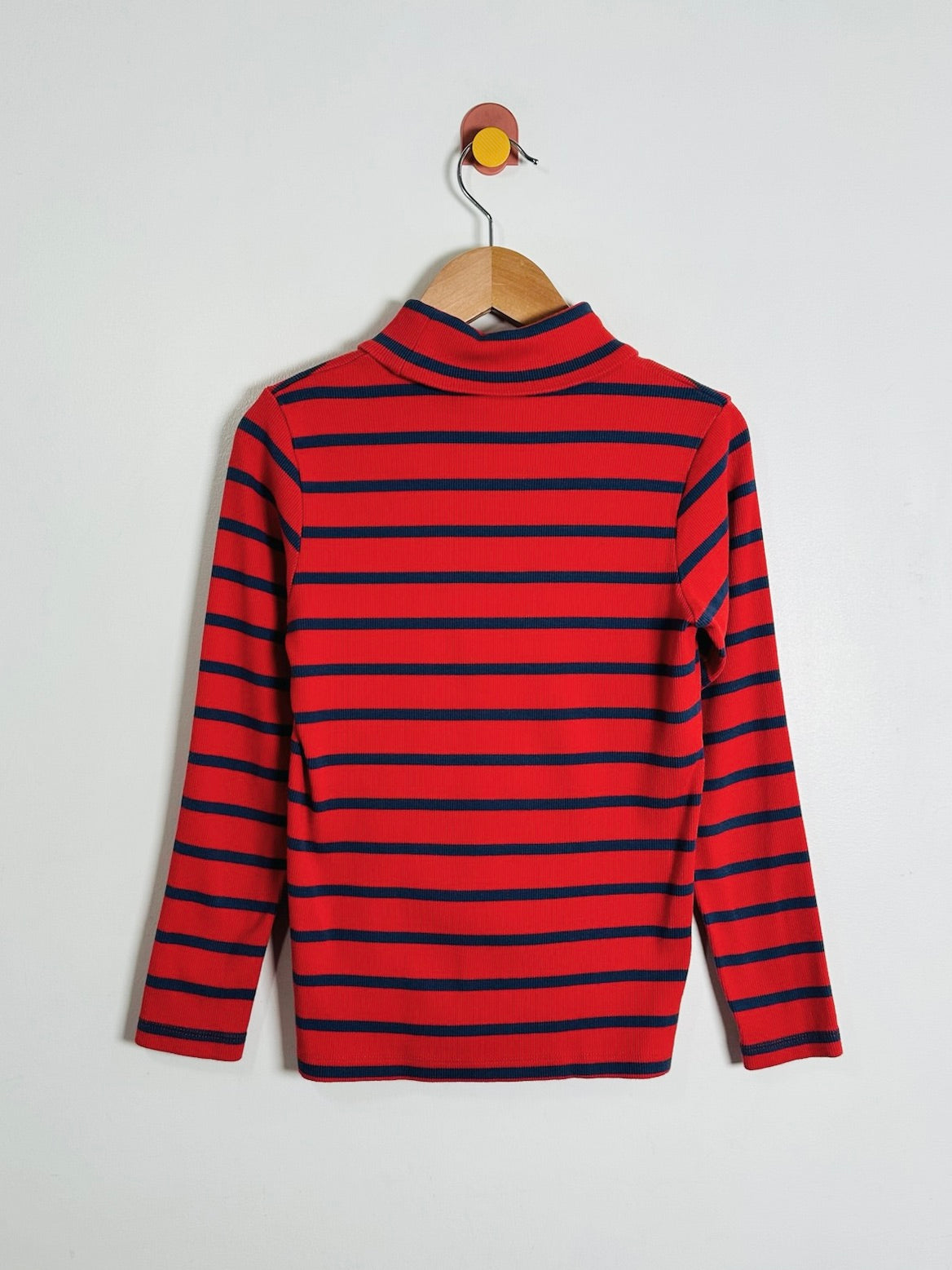 A red and navy blue horizontally striped long-sleeve mock neck sweater hanging on a wooden hanger against a plain light gray wall.