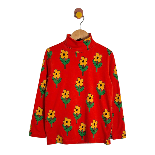 A red long-sleeve mock-neck top with an all-over yellow flower and green leaf pattern, hanging on a wooden hanger against a plain white background.