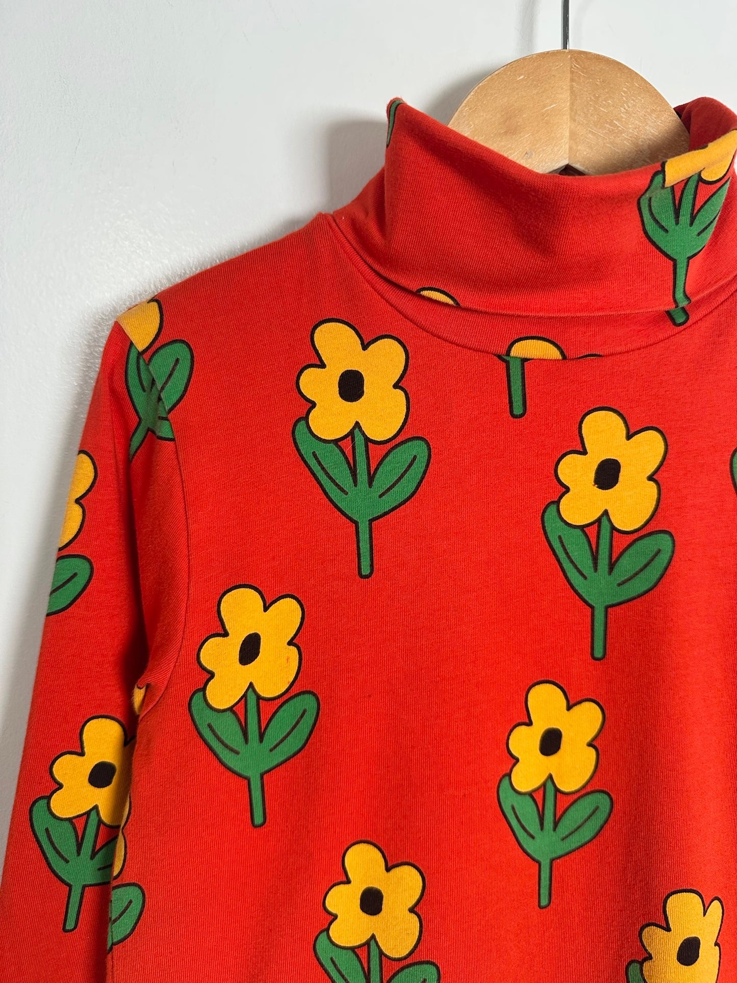 A red mock-neck long-sleeve top featuring a repeating yellow flower and green leaf pattern, shown hanging on a wooden hanger against a plain light gray wall.