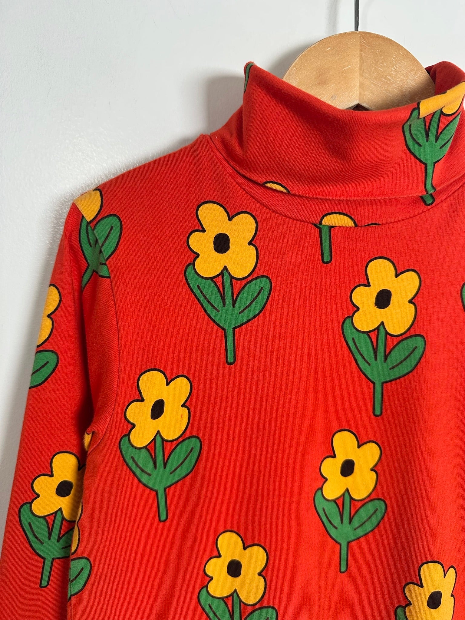 A red mock-neck long-sleeve top featuring a repeating yellow flower and green leaf pattern, shown hanging on a wooden hanger against a plain light gray wall.