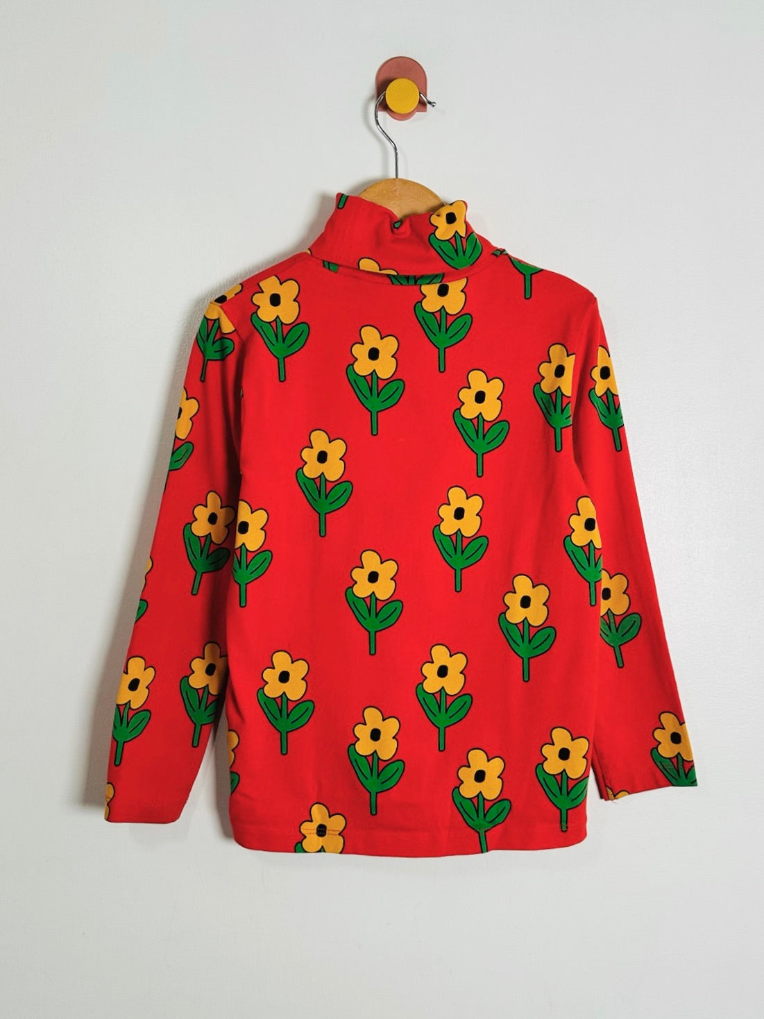 A red long-sleeve mock neck top with a repeating yellow flower and green leaf pattern, hanging on a wooden hanger against a plain light gray wall.