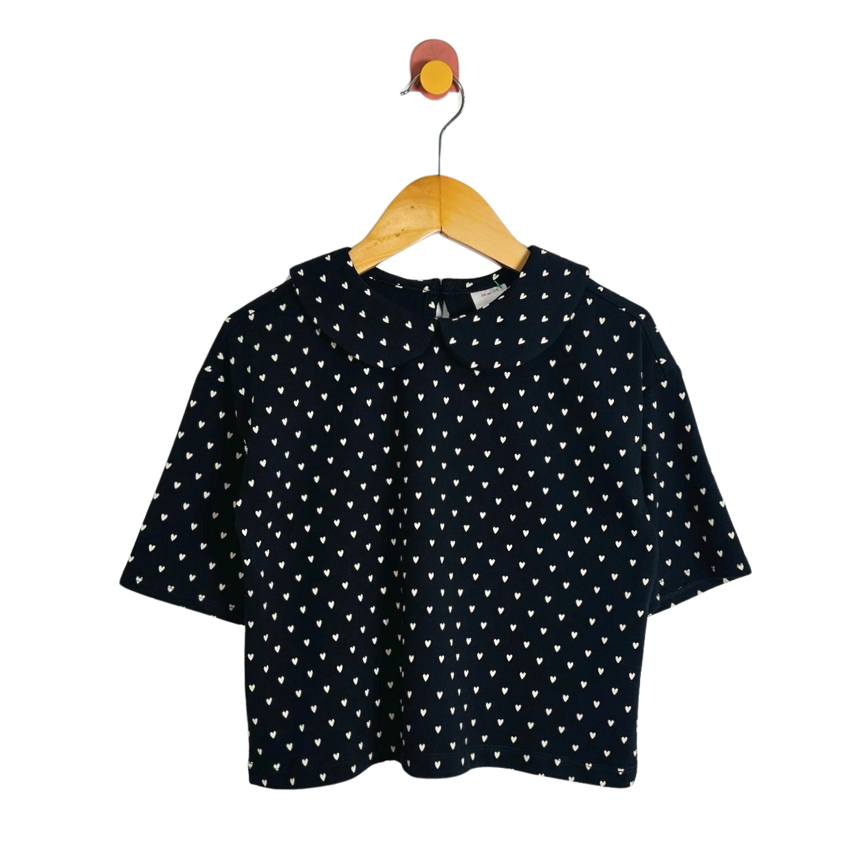 A short-sleeve black top with a white heart pattern, hanging on a wooden hanger with a colorful hook against a plain white background.