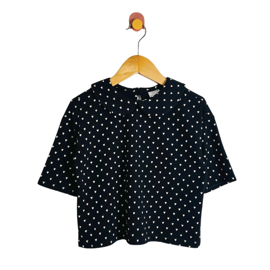 A short-sleeve black top with a white heart pattern, hanging on a wooden hanger with a colorful hook against a plain white background.