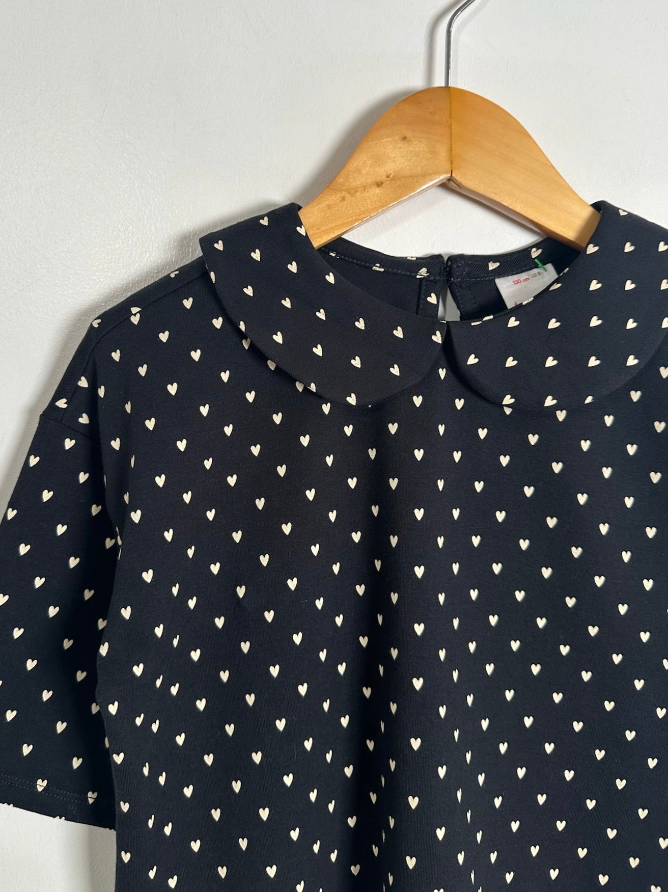 A short-sleeve black top with a repeating small white heart pattern, hanging on a wooden hanger against a plain light gray wall.
