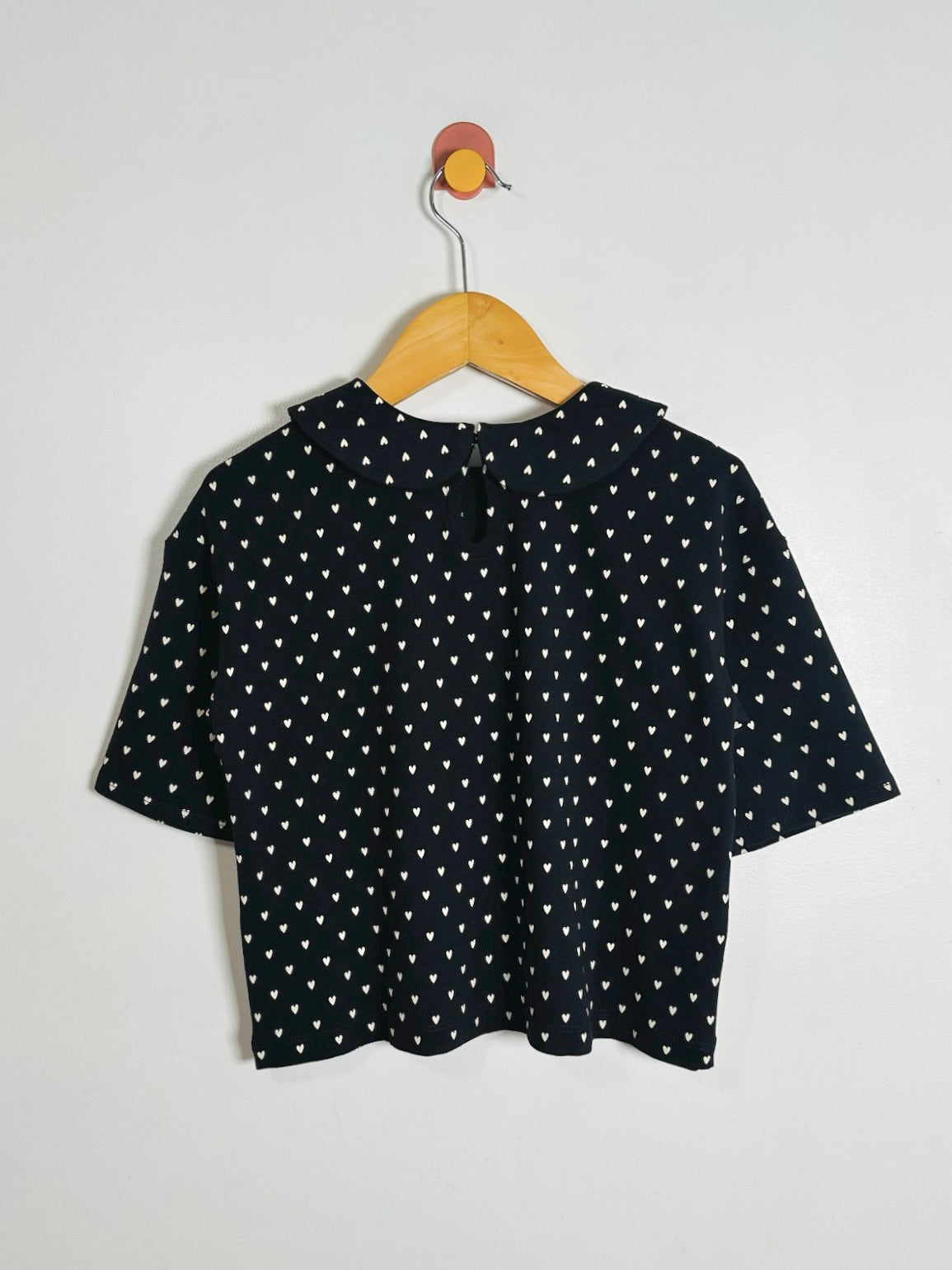 A black short-sleeve top with a white polka-dot pattern, hanging on a wooden hanger against a plain light gray wall.
