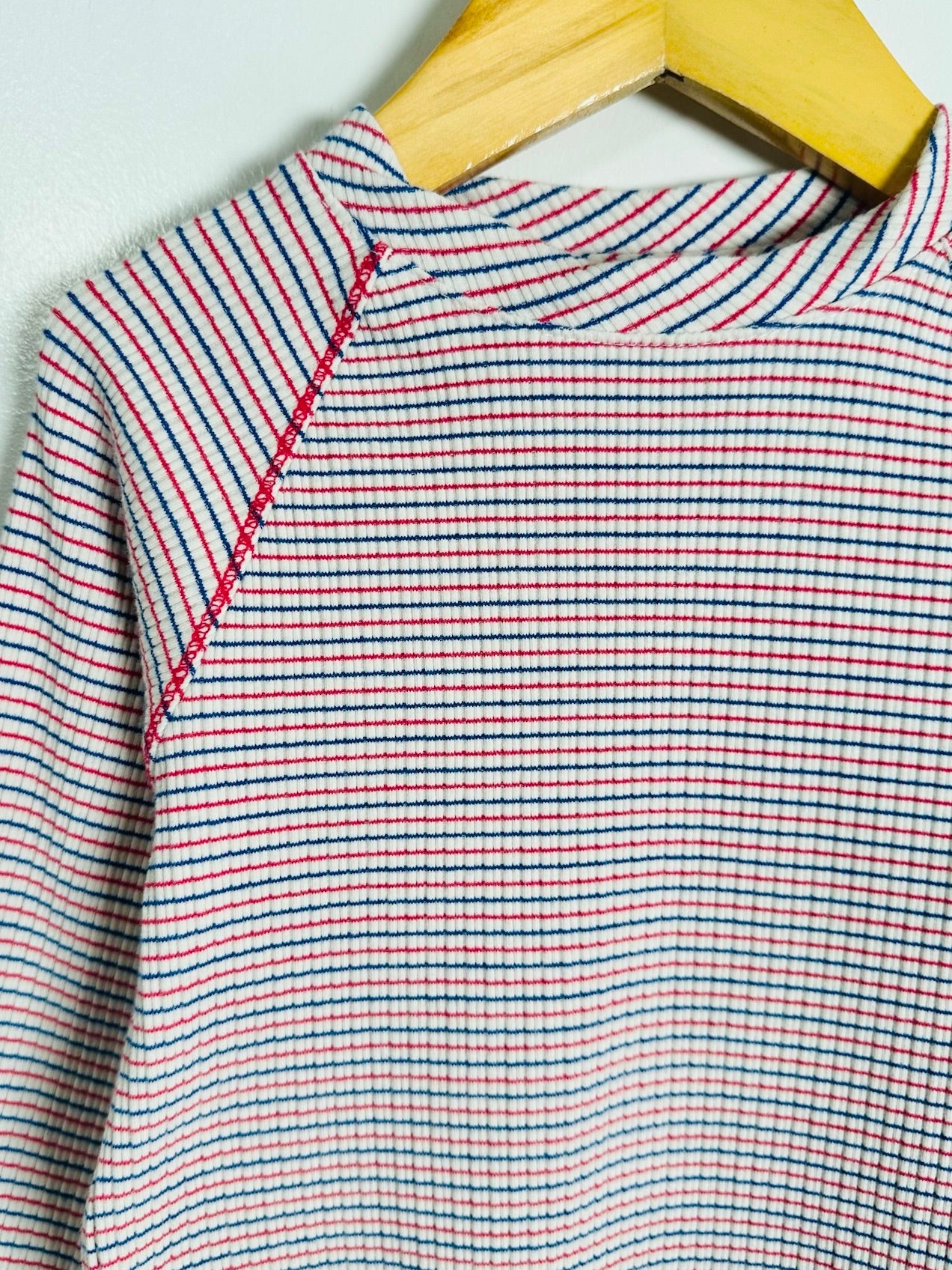 Close-up of a striped knit top hanging on a wooden hanger against a light gray wall. The garment has thin horizontal stripes in red, navy blue, and white, with a visible red contrast stitch along the shoulder seam.